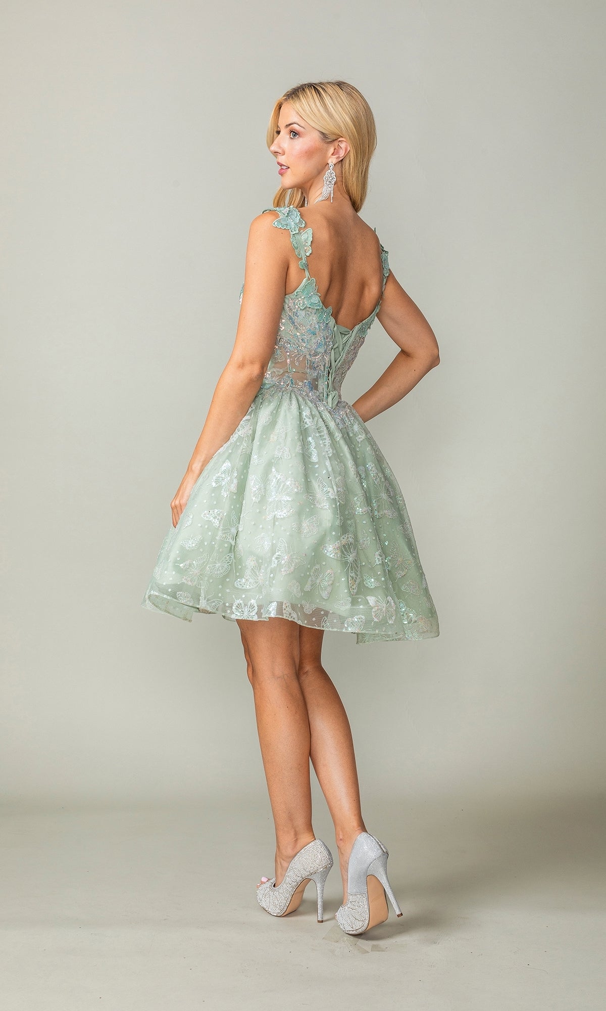 A woman stands facing away, wearing the Sparkly Short Butterfly Homecoming Dress 3361 in light green with lace details, a flared skirt, and silver high heels against a plain background.